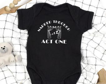 May include: Black baby onesie with the text "NAPPED THROUGH ACT ONE" in white. The design includes a theater curtain and the word "zzz" to represent sleep. The onesie is on a white and black patterned blanket.