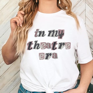 May include: White t-shirt featuring the text "in my theatre era" in a 3D design. The text uses black, grey, and mauve colours. The t-shirt is a casual style, suitable for everyday wear.