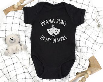 May include: Black baby onesie with the text "DRAMA RUNS IN MY DIAPERS" in white. The onesie features a graphic of two theater masks and a safety pin. The onesie is on a white and black patterned blanket with a teddy bear and wooden beads.