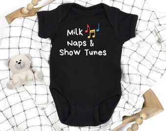 May include: Black baby bodysuit with the text "Milk, Naps & Show Tunes" in white. The design includes musical notes in red, yellow, and blue. The bodysuit is on a white and black patterned blanket with a small stuffed bear.