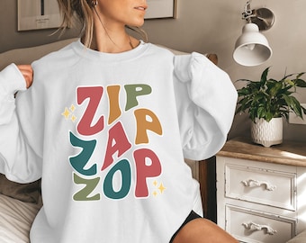May include: White sweatshirt with the words "ZIP ZAP ZOP" in colorful, playful letters. The design includes red, green, orange, and blue text with yellow star accents. A casual, comfortable top.
