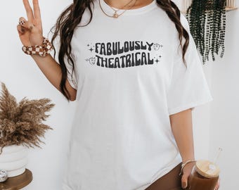 May include: White oversized t-shirt with black text that reads "FABULOUSLY THEATRICAL" in a retro font. The text is surrounded by small stars and stylized faces.