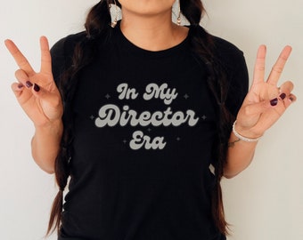 May include: Black t-shirt with the text "In My Director Era" in a retro font. The text is in a light gray color with a white outline. The shirt has a crew neck.