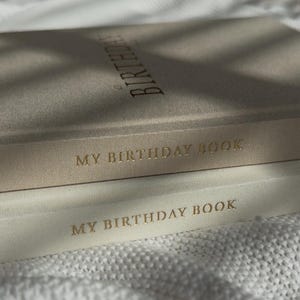 Puede incluir: Primer plano de dos diarios "My Birthday Book". El diario superior es de color beige claro con la palabra "Birthday" impresa en dorado. El diario inferior es de color crema con las palabras "My Birthday Book" impresas en dorado.
