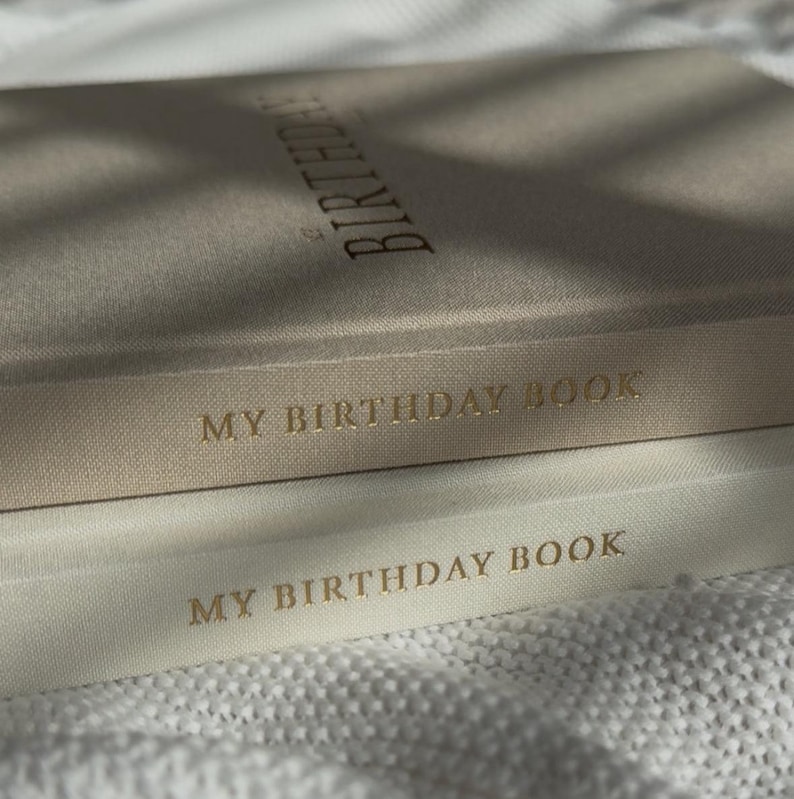 Puede incluir: Primer plano de dos diarios "My Birthday Book". El diario superior es de color beige claro con la palabra "Birthday" impresa en dorado. El diario inferior es de color crema con las palabras "My Birthday Book" impresas en dorado.