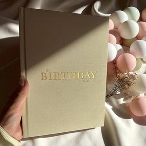 Puede incluir: Un diario beige con el texto dorado "MY BIRTHDAY 2003" en la portada. El diario est&aacute; sostenido por una mano y est&aacute; junto a una colecci&oacute;n de globos blancos y rosas.