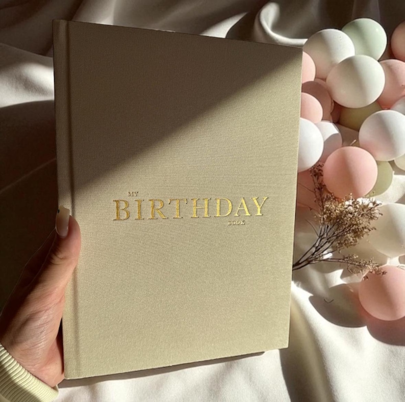 Puede incluir: Un diario beige con el texto dorado "MY BIRTHDAY 2003" en la portada. El diario est&aacute; sostenido por una mano y est&aacute; junto a una colecci&oacute;n de globos blancos y rosas.