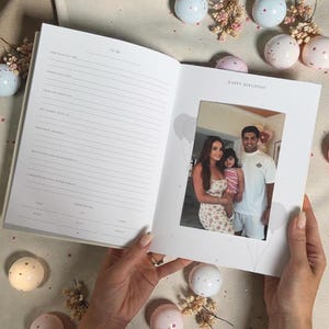 Puede incluir: Un libro de recuerdos crema abierto, con p&aacute;ginas rayadas y un marco de fotos. El marco contiene una foto familiar. Las palabras "Happy Birthday" est&aacute;n impresas encima de la foto. Esferas decorativas y flores rodean el libro.