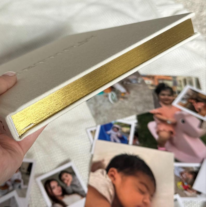 Puede incluir: Un &aacute;lbum de fotos color crema con bordes dorados. El &aacute;lbum est&aacute; abierto, con varias fotograf&iacute;as impresas esparcidas alrededor. La cubierta del &aacute;lbum tiene una textura sutil y un dise&ntilde;o minimalista, ideal para preservar recuerdos.