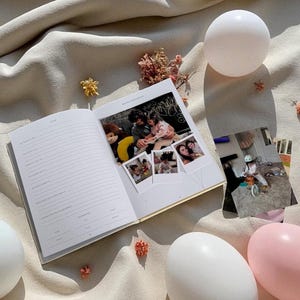 Puede incluir: Un diario abierto con p&aacute;ginas rayadas y un borde dorado, con fotograf&iacute;as y las palabras "Happy Birthday". El diario est&aacute; rodeado de elementos decorativos, incluyendo adornos blancos y rosas en forma de huevo y flores secas, todo sobre una tela beige.
