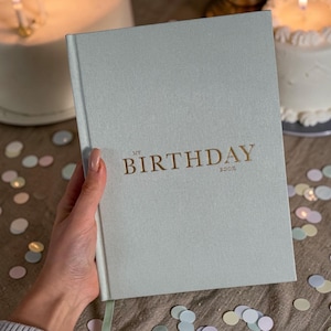 Puede incluir: Un libro verde claro "My Birthday Book" con letras doradas. El libro est&aacute; en la mano de una persona. El libro est&aacute; sobre una superficie marr&oacute;n con confeti y dos pasteles en el fondo.