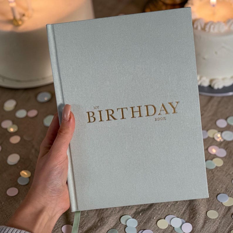 Puede incluir: Un libro verde claro "My Birthday Book" con letras doradas. El libro est&aacute; en la mano de una persona. El libro est&aacute; sobre una superficie marr&oacute;n con confeti y dos pasteles en el fondo.
