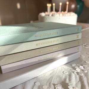 Puede incluir: Una pila de diarios "My Birthday Book" de colores pastel con letras doradas. Los libros son en tonos de azul, verde, beige y lavanda. Un pastel de cumplea&ntilde;os blanco con velas encendidas est&aacute; en el fondo, junto con confeti esparcido.