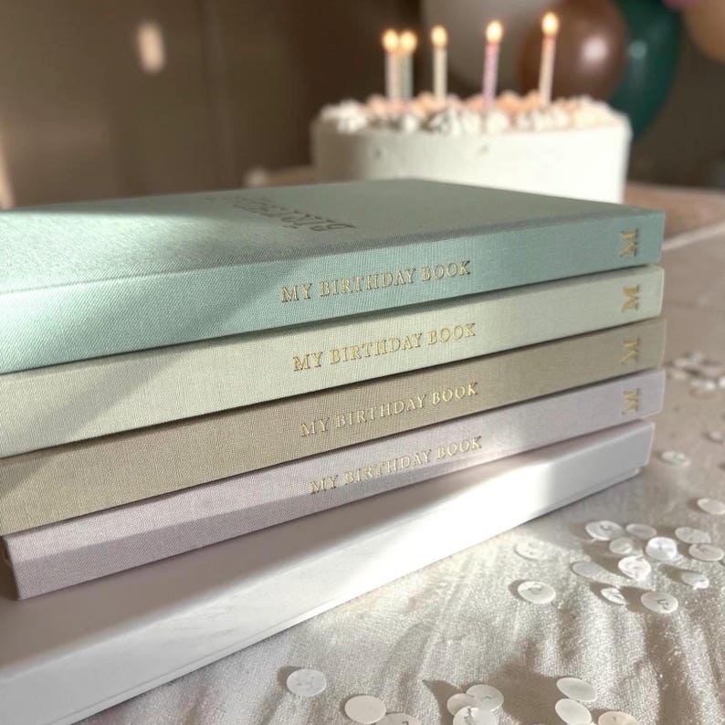 Puede incluir: Una pila de diarios "My Birthday Book" de colores pastel con letras doradas. Los libros son en tonos de azul, verde, beige y lavanda. Un pastel de cumplea&ntilde;os blanco con velas encendidas est&aacute; en el fondo, junto con confeti esparcido.