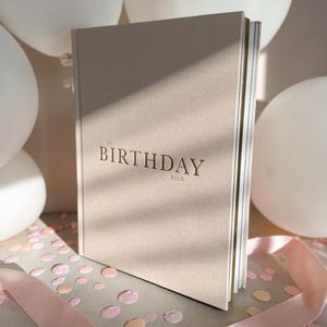 Puede incluir: Un libro beige de tapa dura titulado "My Birthday Book" en una fuente serif. El libro est&aacute; ligeramente inclinado, revelando las p&aacute;ginas doradas. Globos blancos, confeti y una cinta rosa en el fondo.