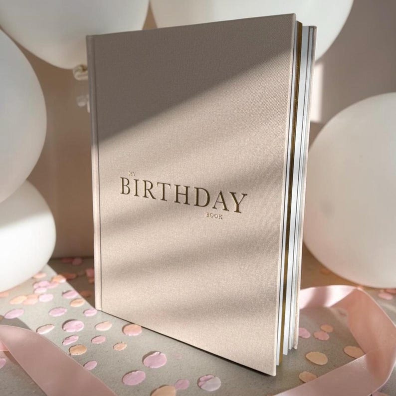 Puede incluir: Un libro beige de tapa dura titulado "My Birthday Book" en una fuente serif. El libro est&aacute; ligeramente inclinado, revelando las p&aacute;ginas doradas. Globos blancos, confeti y una cinta rosa en el fondo.