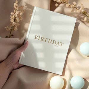 Puede incluir: Un libro crema "My Birthday Book" con letras doradas est&aacute; abierto. El libro est&aacute; sobre una tela suave de color melocot&oacute;n, con ramitas de flores secas y dos esferas azul claro. La portada del libro presenta las palabras "My Birthday Book".