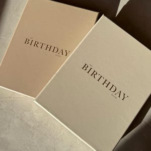 Puede incluir: Dos libros de tapa dura con el texto "MY BIRTHDAY BOOK" en una fuente serif. Un libro es de color beige claro, y el otro es de un beige ligeramente m&aacute;s oscuro. Los libros son rectangulares y parecen diarios o cuadernos.