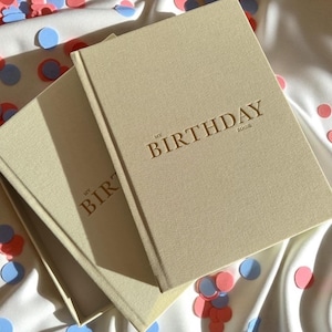 May include: Two beige hardcover books with the gold text "MY BIRTHDAY" and "2008" on the cover. The books are on a white surface with scattered confetti in shades of red, blue, and pink.