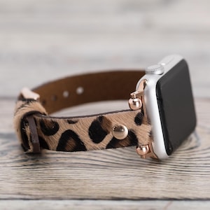May include: A silver Apple Watch with a brown and black leopard print leather band. The band has rose gold accents and a buckle.