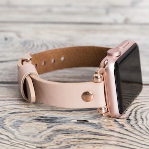 May include: A rose gold Apple Watch with a light pink leather band. The band has a rose gold buckle and a rose gold connector.