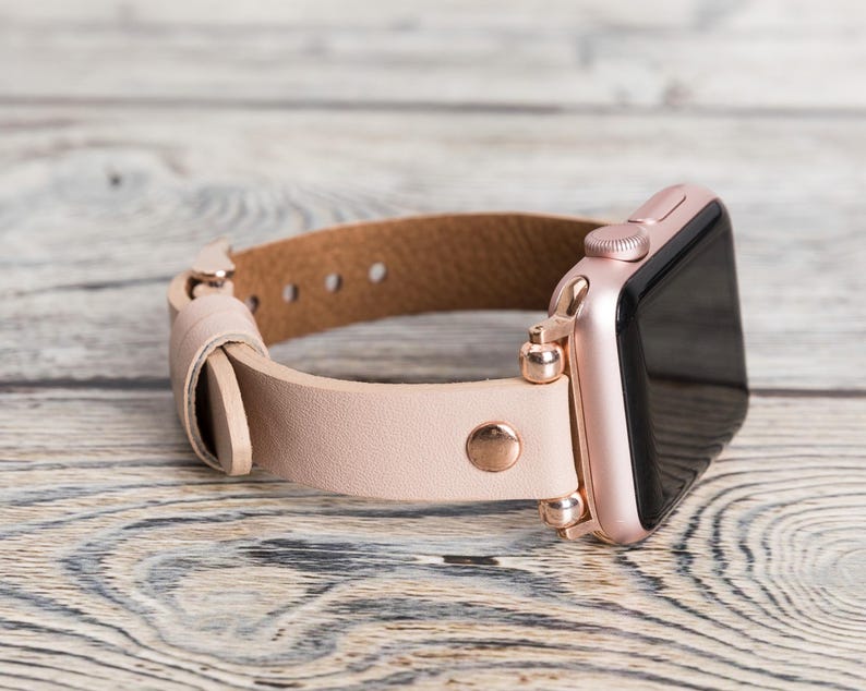 May include: A rose gold Apple Watch with a light pink leather band. The band has a rose gold buckle and a rose gold connector.