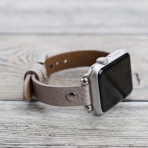 May include: A silver Apple Watch with a light gray leather band. The band has a textured finish and silver metal accents.