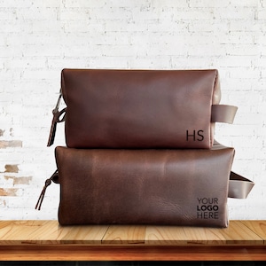 May include: Two brown leather travel bags stacked on top of each other. The top bag has the initials "HS" embossed on the front. The bottom bag has the text "YOUR LOGO HERE" embossed on the front.