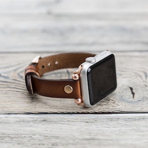 Op de afbeelding: Een bruin leren horlogebandje met ros&eacute;gouden accenten en een zilveren Apple Watch.