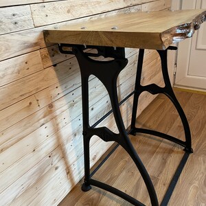 May include: A wooden table top with a live edge sits on top of black metal industrial style legs. The table is mounted to a wall.