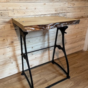 May include: A wooden table top with live edge details sits on a black metal industrial style base. The table is made of light wood with a natural finish. The base is made of cast iron and has a black finish.