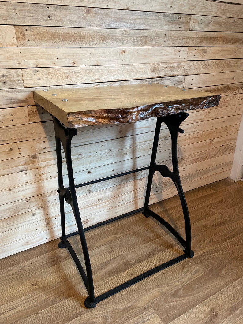 May include: A wooden table top with live edge details sits on a black metal industrial style base. The table is made of light wood with a natural finish. The base is made of cast iron and has a black finish.