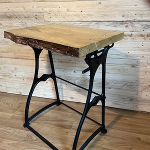 May include: A wooden table top with a live edge sits on a black metal adjustable base. The table top is made of light wood and has a natural finish. The base is made of cast iron and has a black finish.