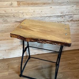 May include: A wooden table top with a live edge and a black metal base. The table top is made of light wood and has a natural finish. The base is made of sturdy metal and has a black finish.