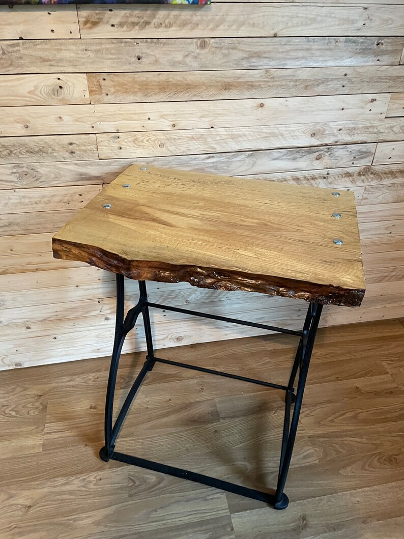 May include: A wooden table top with a live edge and a black metal base. The table top is made of light wood and has a natural finish. The base is made of sturdy metal and has a black finish.