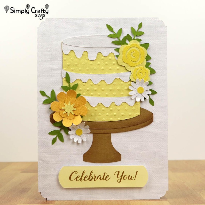 May include: A handmade greeting card featuring a yellow layered cake with white frosting and yellow flowers. The cake sits on a brown cake stand, and the card has the text "Celebrate You!" on a yellow banner.
