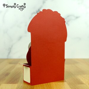 May include: Red card with a pop-up design. The card is folded and has a small white square on the front. The card is on a wooden surface.