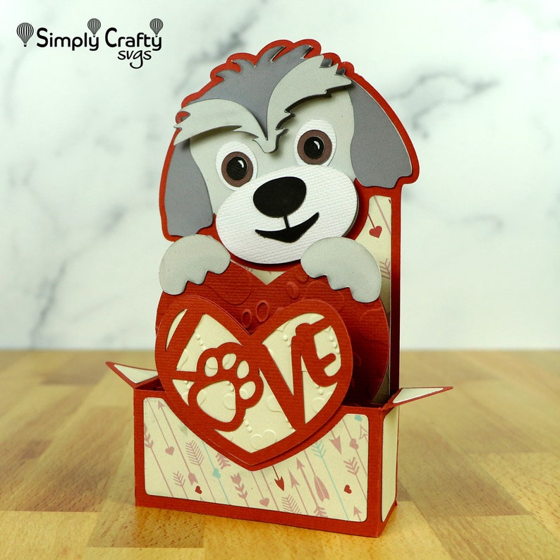 May include: A grey and white dog holding a red heart with the word "LOVE" and a paw print inside. The heart is in a red box with a white and red patterned background.