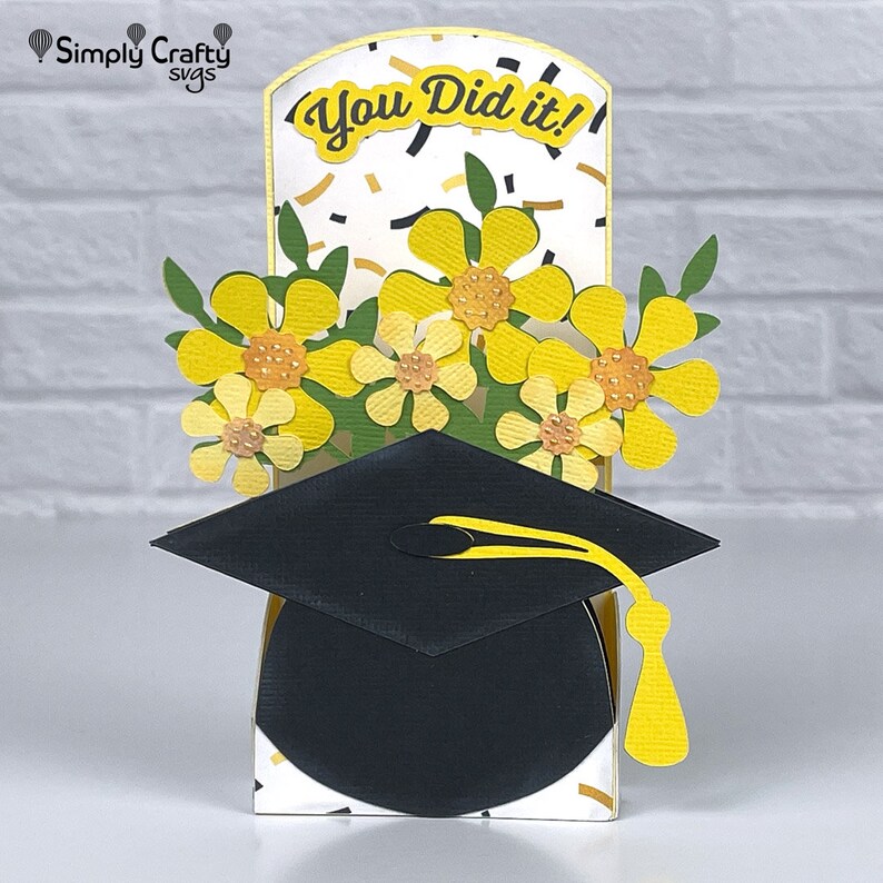Op de afbeelding: Een afstudeerkaart met een pop-up afstudeerhoed en gele bloemen. De kaart heeft een witte achtergrond met gouden confetti en de tekst "You Did It!" op de voorkant.