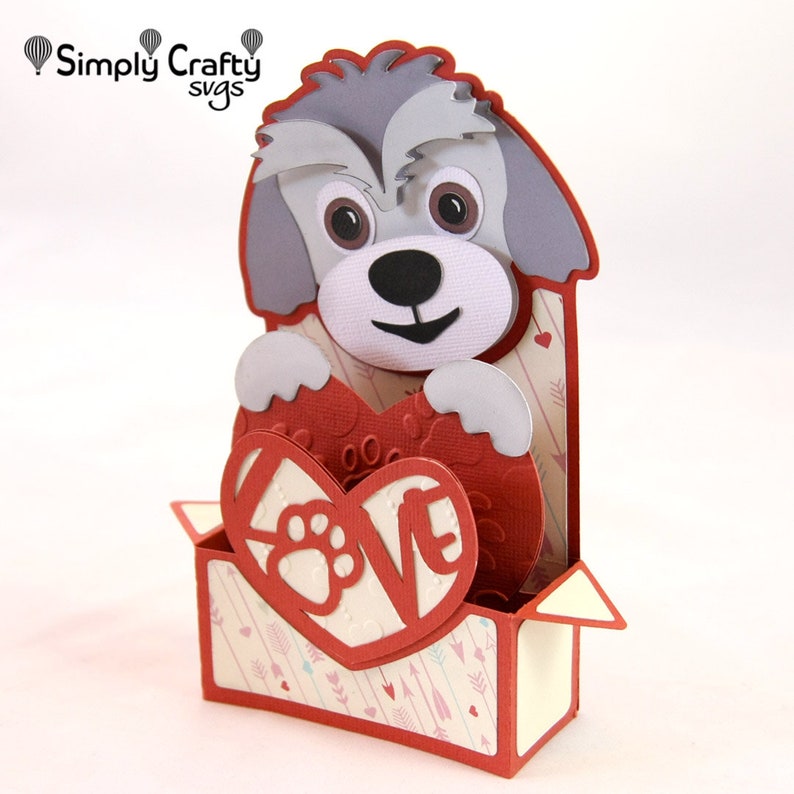 May include: A grey and white dog holding a red heart with the word "LOVE" cut out and a paw print inside. The heart is in a red box with a white and pink patterned background.