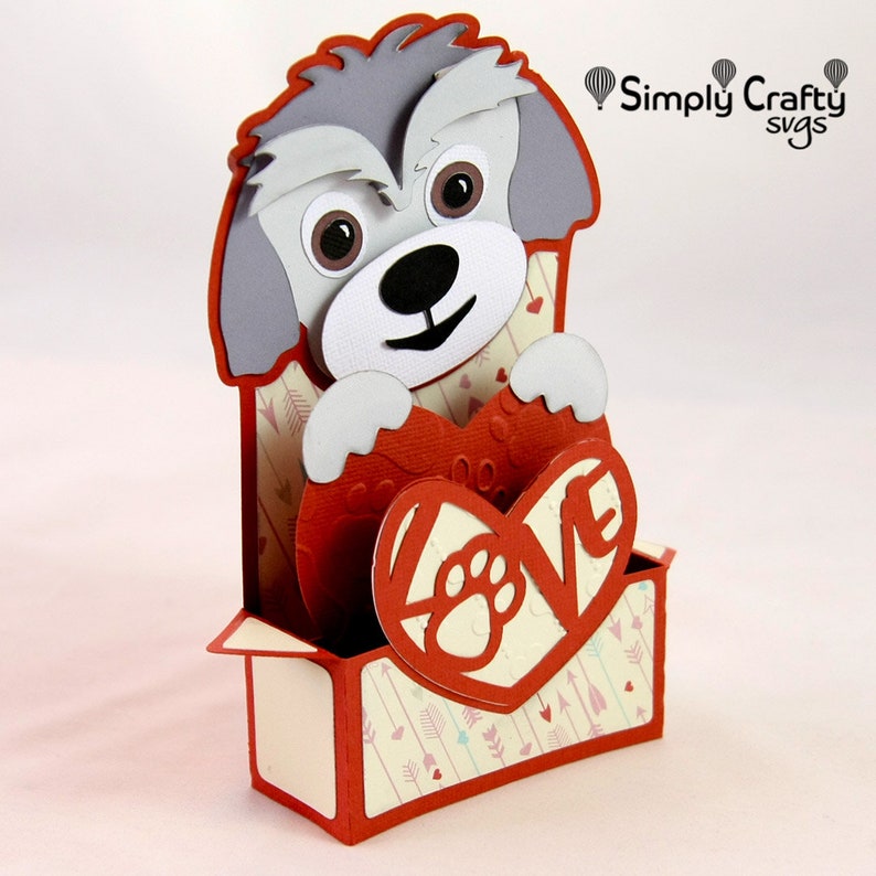 May include: A grey and white dog holding a red heart with the word "LOVE" cut out. The heart is in a box with a red and white pattern of arrows and hearts.