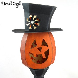 May include: A papercraft pumpkin jack-o'-lantern with a black top hat and a white and black pinwheel on top. The pumpkin has a carved face with a wide smile and a small nose.