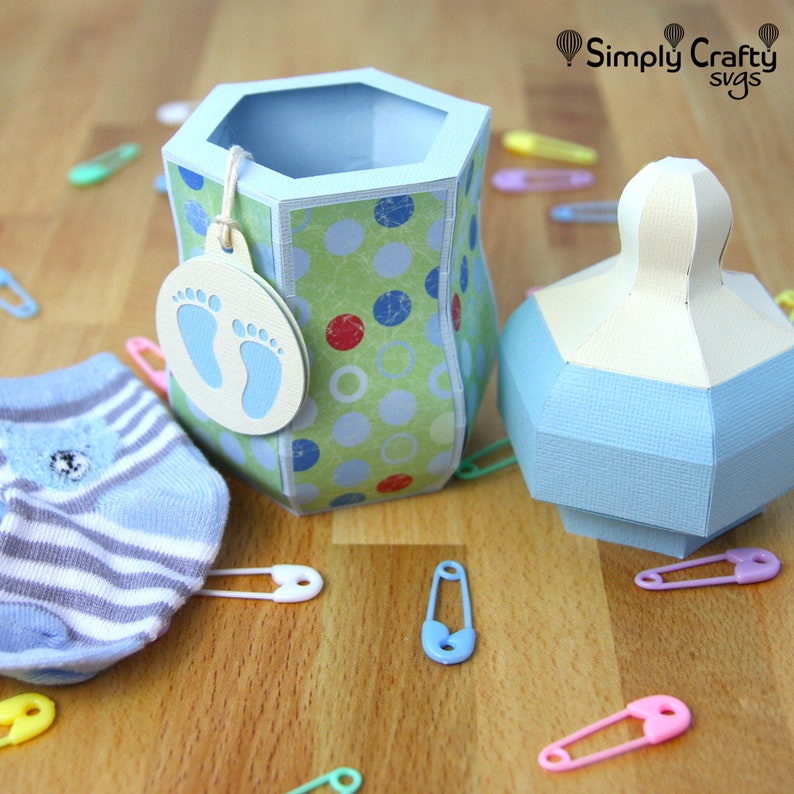 Puede incluir: Una caja de regalo con forma de biber&oacute;n azul y verde con una etiqueta blanca que dice "Simply Crafty SVGs". La caja est&aacute; decorada con lunares y tiene un calcet&iacute;n a rayas azul y blanco a su lado.