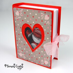 May include: A red and gray Valentine's Day-themed gift box with a heart-shaped cutout revealing chocolate candies. The box is decorated with pink polka dots and heart-shaped accents. A sheer pink ribbon is tied around the side.