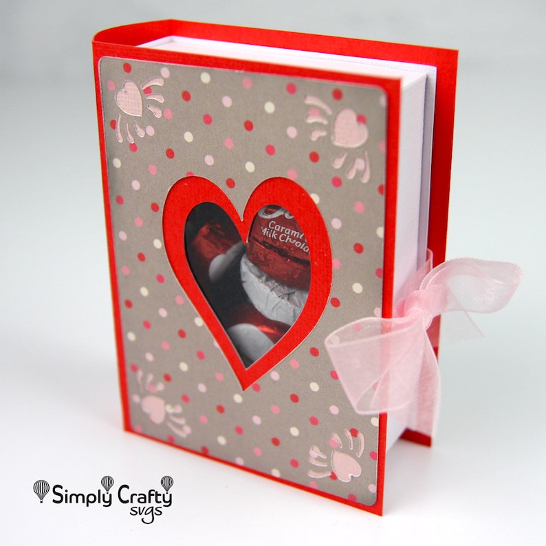 May include: A red and gray Valentine's Day-themed gift box with a heart-shaped cutout revealing chocolate candies. The box is decorated with pink polka dots and heart-shaped accents. A sheer pink ribbon is tied around the side.