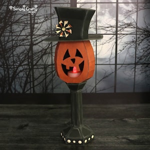 May include: A papercraft jack-o'-lantern with a black top hat and a lit candle inside. The jack-o'-lantern is on a black pedestal with white dots. The background is a blurry image of a window with a moon and trees.