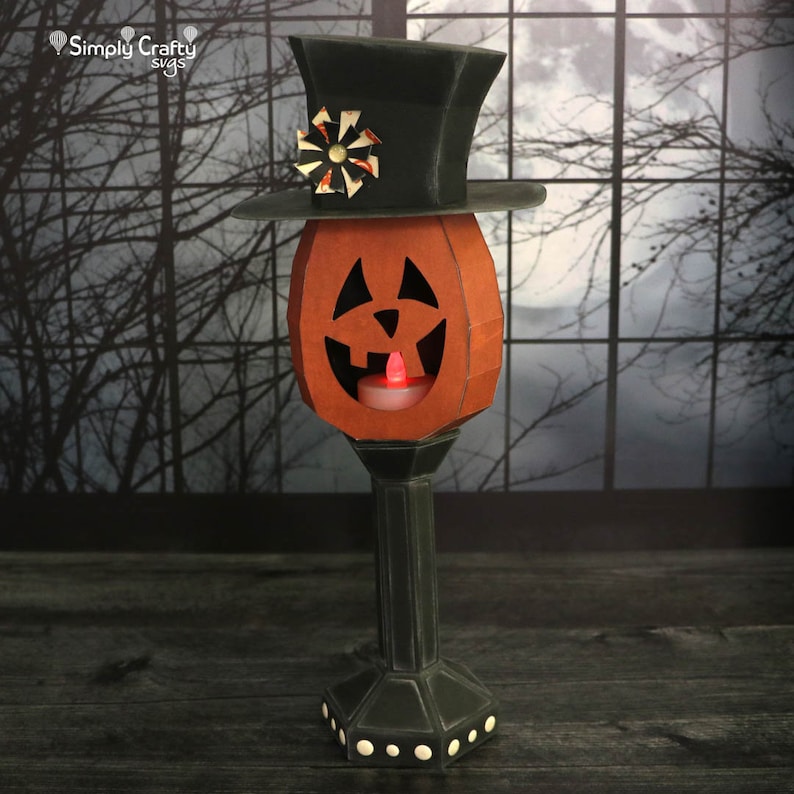 May include: A papercraft jack-o'-lantern with a black top hat and a lit candle inside. The jack-o'-lantern is on a black pedestal with white dots. The background is a blurry image of a window with a moon and trees.