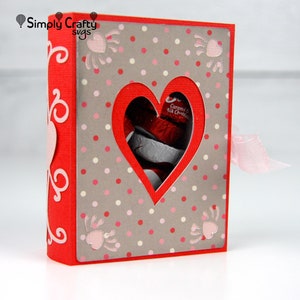 May include: A red paper gift box with a heart-shaped window revealing wrapped chocolates. The box has a gray polka-dot pattern with pink accents and a pink ribbon. The text "Simply Crafty svgs" is in the upper left corner.