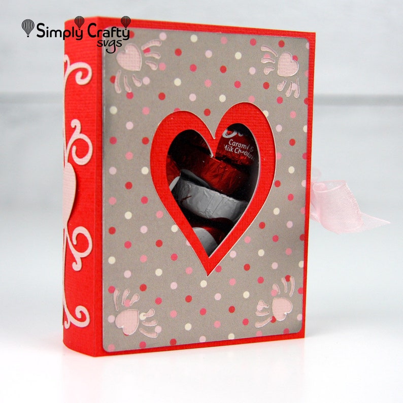 May include: A red paper gift box with a heart-shaped window revealing wrapped chocolates. The box has a gray polka-dot pattern with pink accents and a pink ribbon. The text "Simply Crafty svgs" is in the upper left corner.