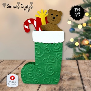 Puede incluir: Un calcetín navideño verde con un puño blanco festoneado, lleno de un oso de peluche marrón, un regalo amarillo y un bastón de caramelo rojo y blanco. La imagen incluye el texto "Simply Crafty Svgs" y "SVG Cut File".